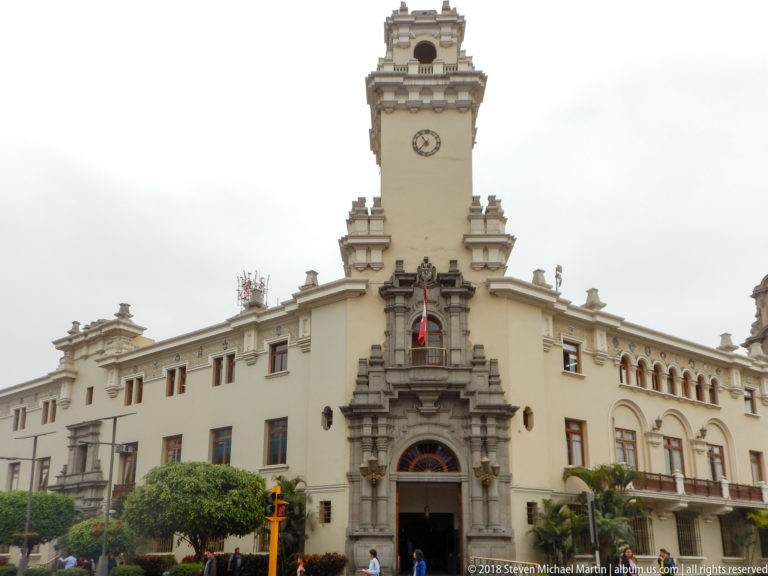 Oblique view of the Municipal Palace of Miraflores with clock tower, Peruvian flag, arched entrance, trees, and pedestrians.