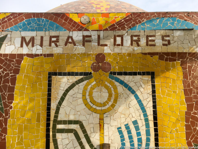 Close view of a yellow broken-tile mosaic reading MIRAFLORES above a stylized multicolored civic emblem.