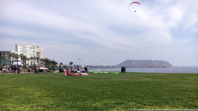 Wide grassy Miraflores clifftop park with paraglider canopies on the ground and in the sky, Pacific Ocean and headland beyond.