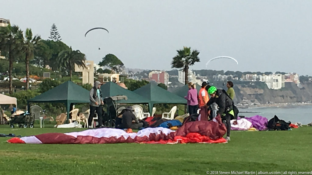 Colorful paraglider canopies spread across a grassy Miraflores park as people gather nearby and several gliders fly beyond the palms.