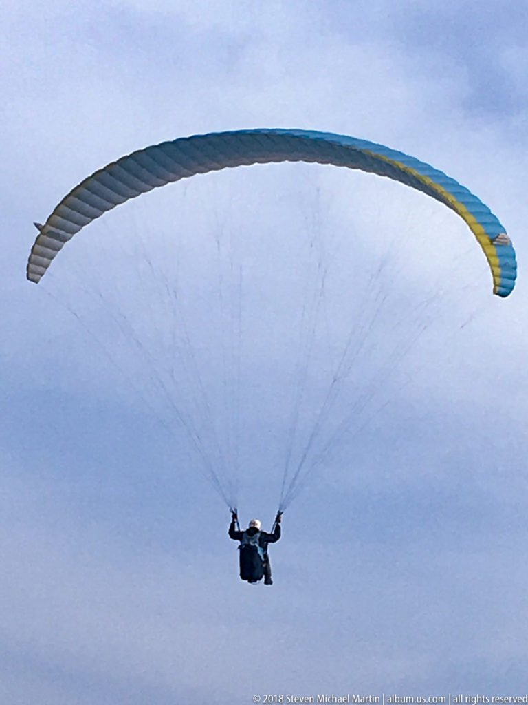 Blue-and-yellow paraglider viewed from below, with the pilot centered beneath a fan of suspension lines against the sky.