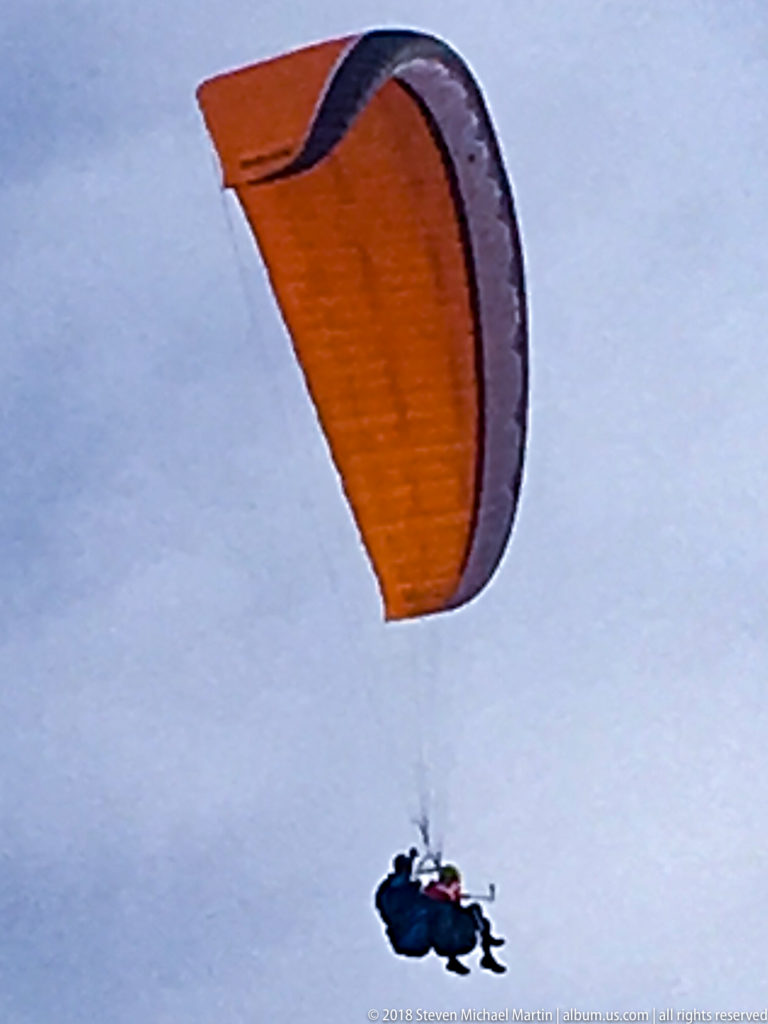 Orange paraglider canopy with a pilot suspended beneath it against a pale overcast sky.