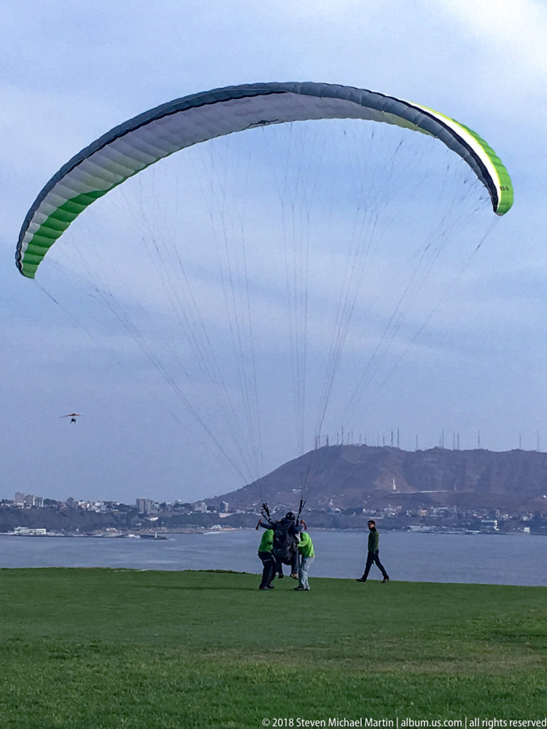 Large green-and-white paraglider canopy descending over a grassy Miraflores park with people, Pacific water, and headland behind.