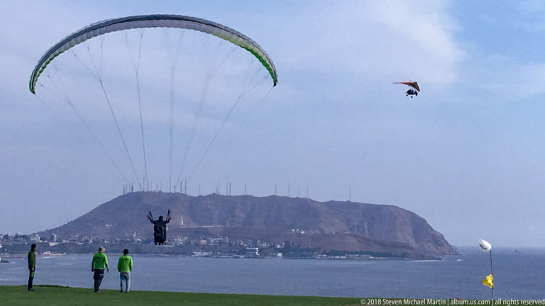 Green-and-white paraglider descending toward a grassy clifftop park, with spectators, another glider, Pacific water, and a distant headland.