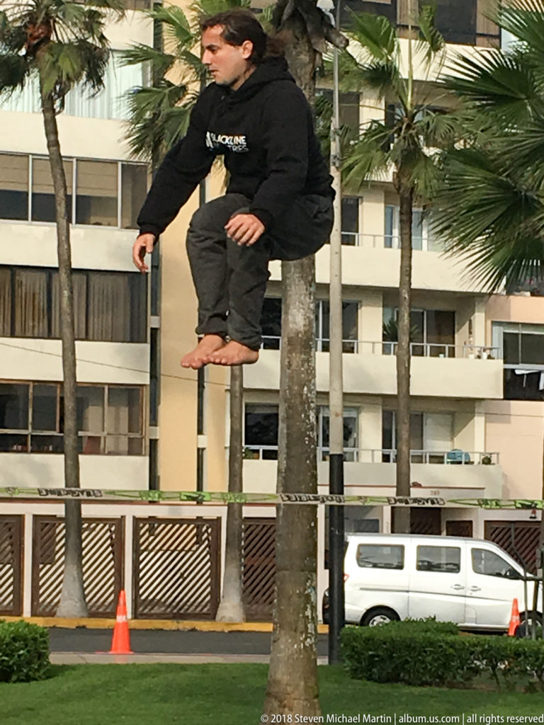 Barefoot paraglider pilot suspended just above a grassy Miraflores lawn, with palm trees and apartment buildings behind him.