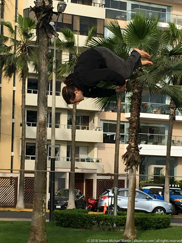 Paraglider pilot flying very low and nearly horizontal between palm trees above a grassy urban park in Miraflores.