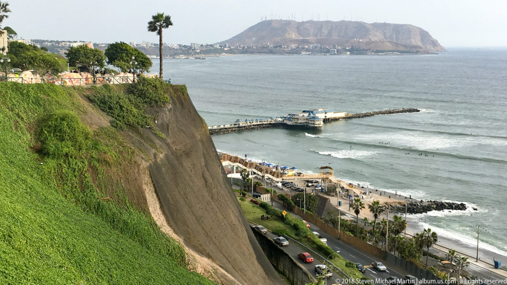 Green Miraflores cliff above the Pacific, with a pier, coastal road and beach below and a broad headland across the water.