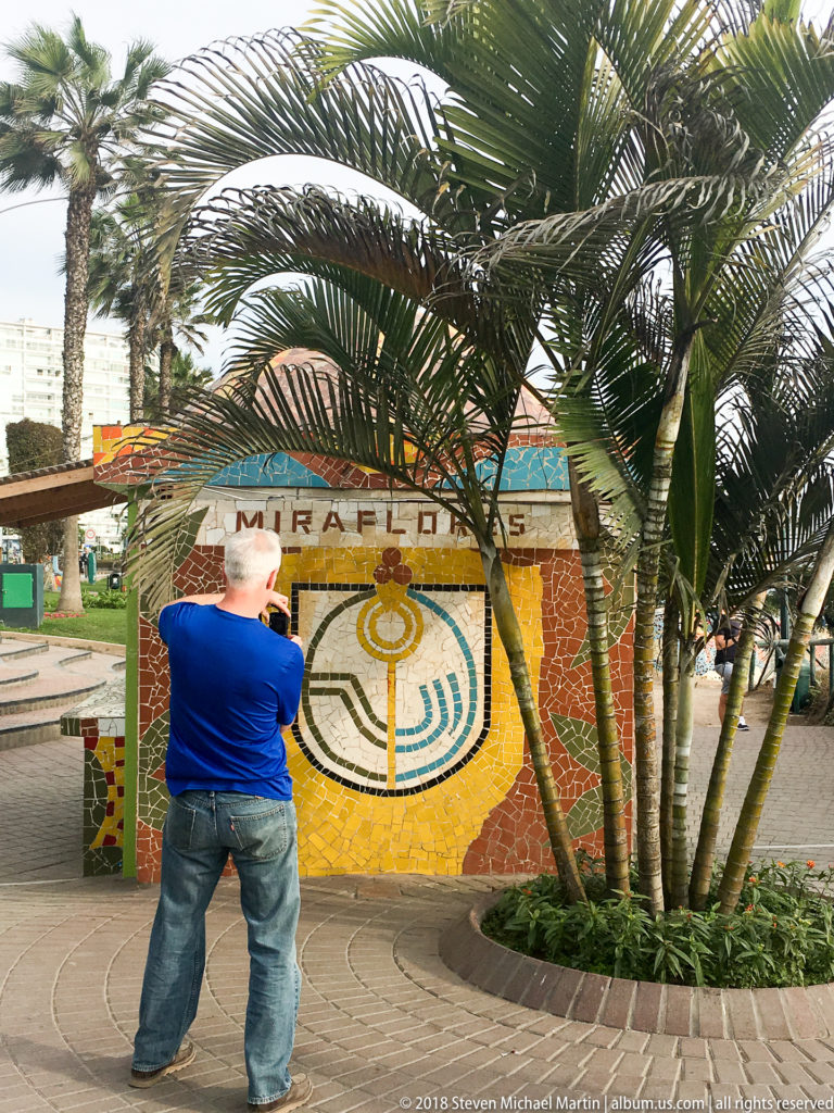 Man photographing a MIRAFLORES mosaic marker partly framed by palm trunks in a landscaped pedestrian area.