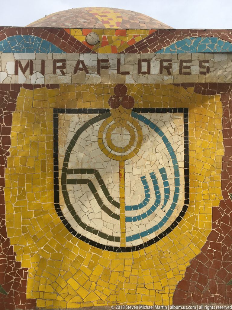 Frontal yellow mosaic panel reading MIRAFLORES above a stylized multicolored civic emblem bordered in dark tile.