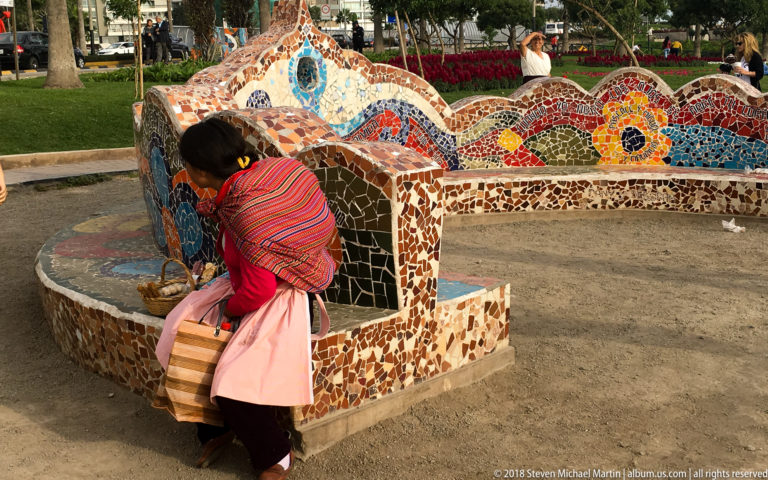 Woman in a striped red garment beside layered, curving multicolored mosaic benches in landscaped Parque del Amor.