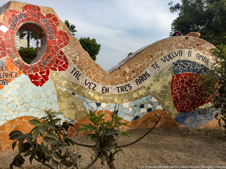 Curving cream mosaic wall with black Spanish lettering, a red heart-like motif, and circular opening amid park landscaping.