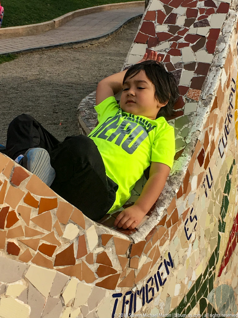 Child in a fluorescent green shirt reclining within a curved multicolored mosaic bench at Parque del Amor.