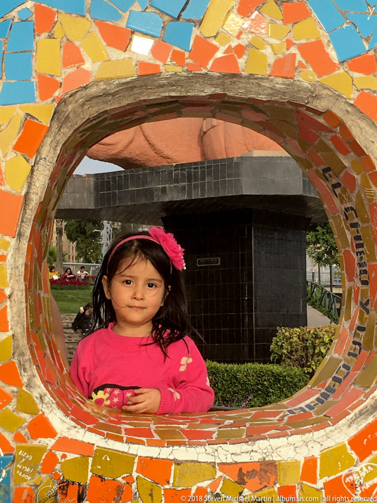 Young child in a pink top looking through a circular opening bordered by colorful broken-tile mosaic at Parque del Amor.