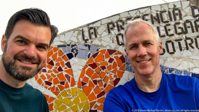 Close portrait of two men beside an orange-and-yellow flower mosaic and black Spanish lettering at Parque del Amor.