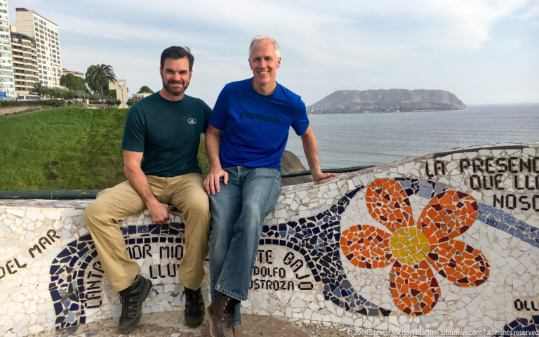 Two men pose beside an orange-flower mosaic wall with Spanish lettering at Parque del Amor, overlooking the Pacific coast.