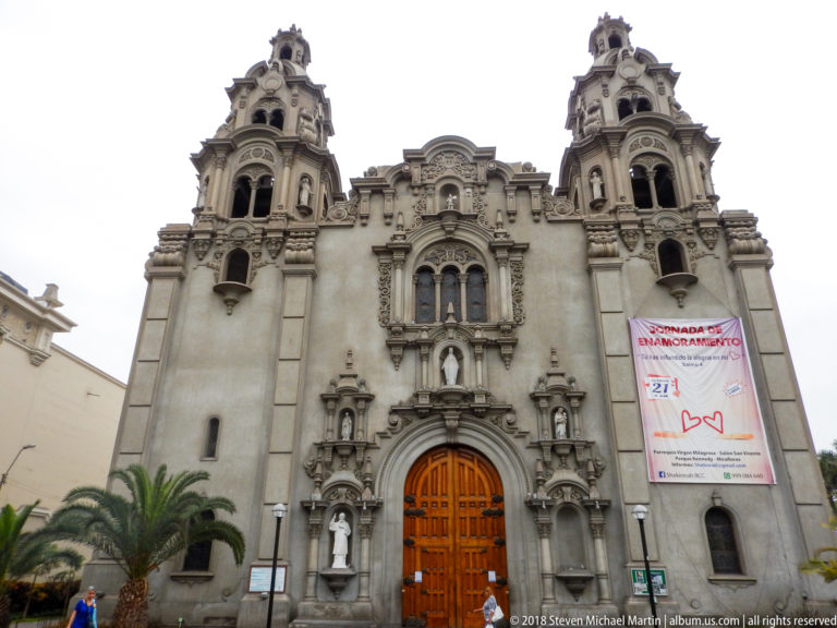 Front view of Iglesia de la Virgen Milagrosa with twin towers, ornate central portal, palms, pedestrians, and a hanging banner.