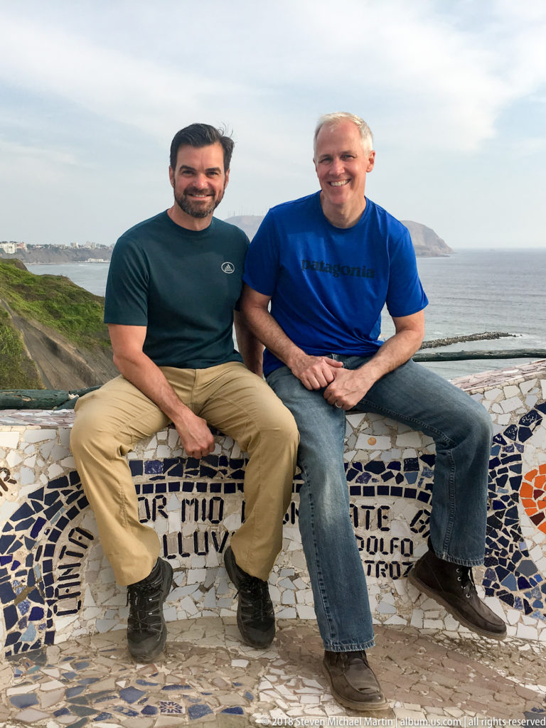 Two men seated on a curving mosaic bench at Parque del Amor, with the Pacific Ocean and distant headland behind them.