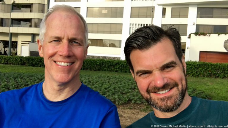Two smiling men pose in a landscaped Miraflores park with green groundcover and a white residential building behind them.
