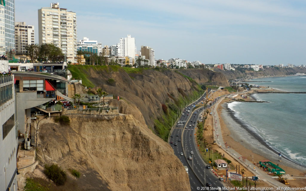 Miraflores high-rises above steep coastal cliffs, with the Costa Verde road, beach, breakwaters, and Pacific Ocean below.