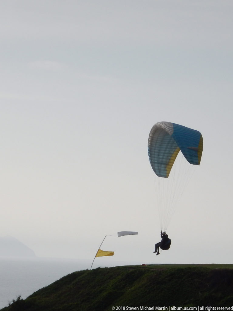 Blue-gray paraglider flying just above a grassy Miraflores clifftop, with a windsock and distant canopy under a pale sky.