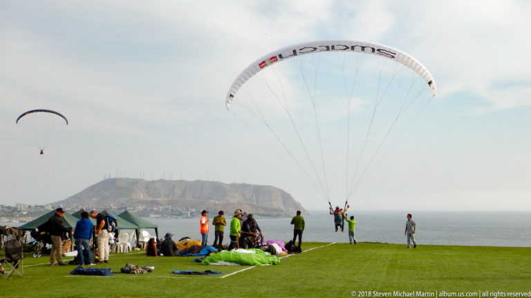 Two paragliders above a grassy Miraflores cliff launch, with people, laid-out wings, the Pacific, and Morro Solar in the distance.