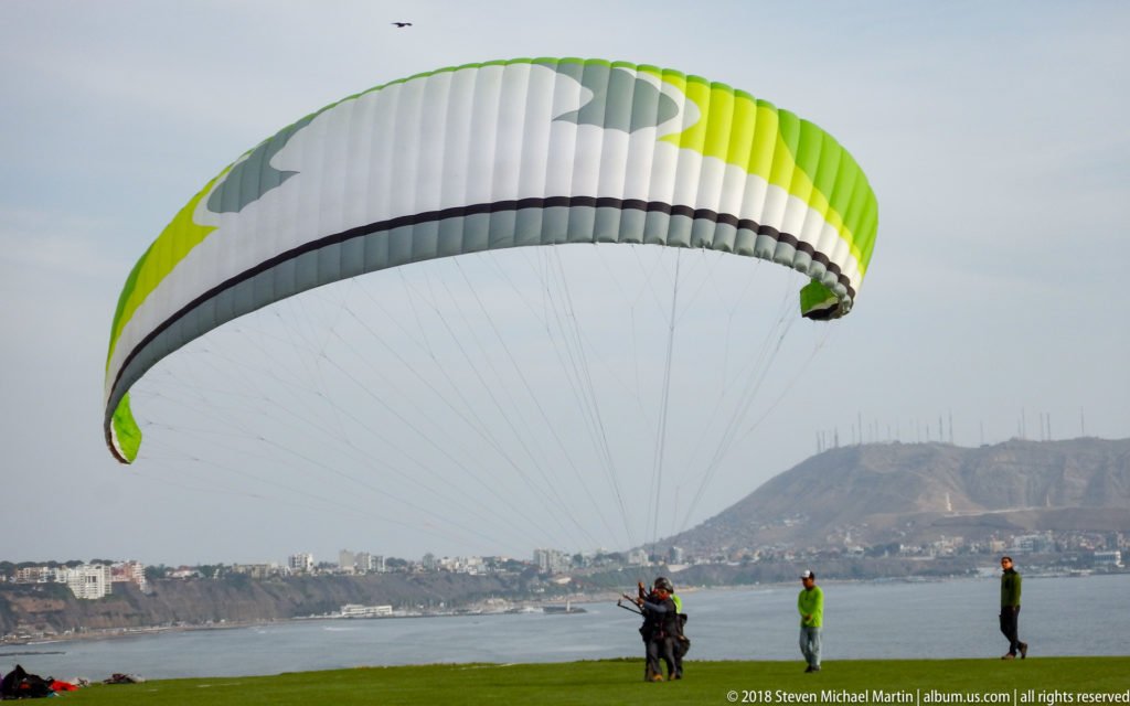 Large green-and-white paraglider canopy inflated above a grassy Miraflores cliff launch, with people and the distant coast behind.