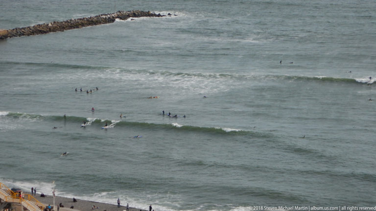 Surfers spread across several breaking Pacific waves below Miraflores, with rocky shoreline visible near the top of the frame.