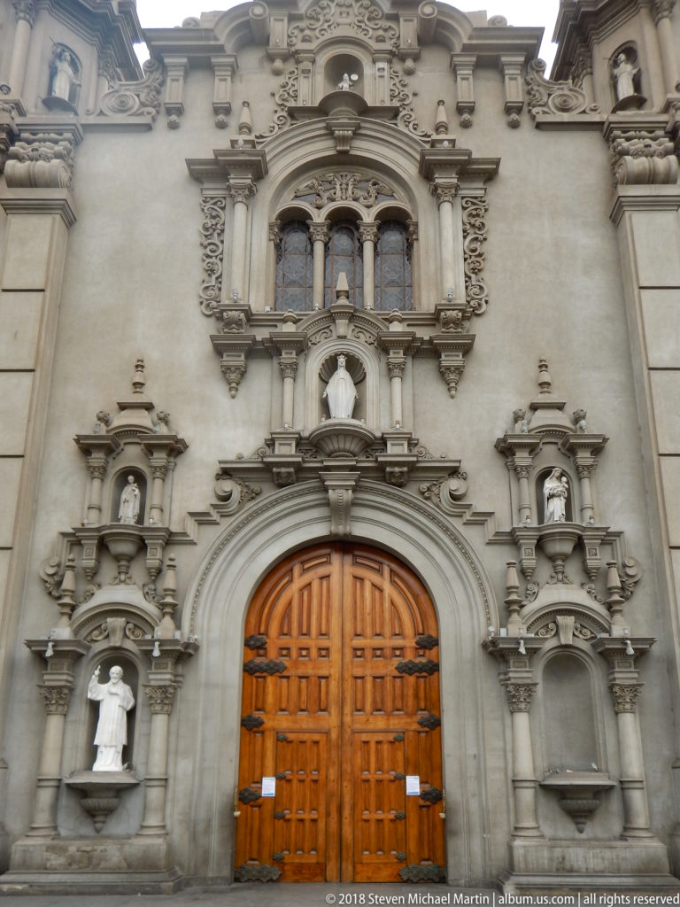 Elaborate church portal with wooden double doors, sculpted saint niches, carved stone ornament, and a triple window above.