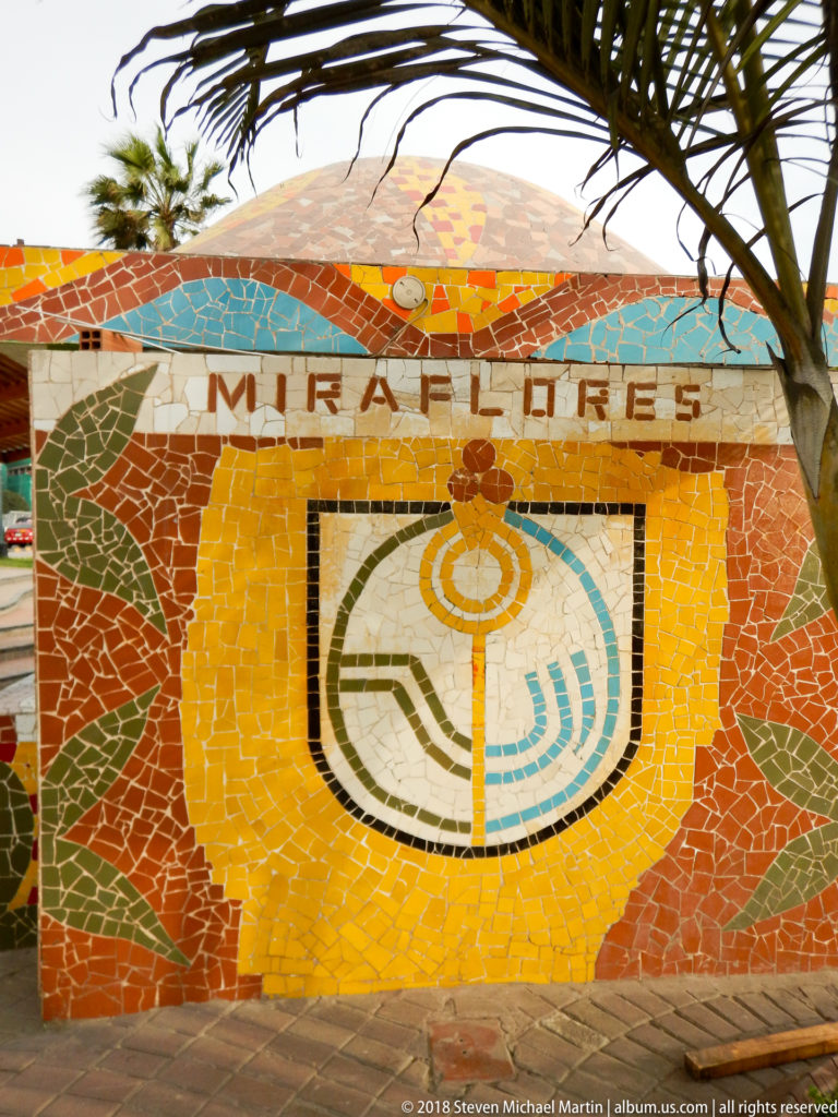 Mosaic panel spelling MIRAFLORES above a centered green-and-blue geometric emblem on a yellow and orange tiled wall.