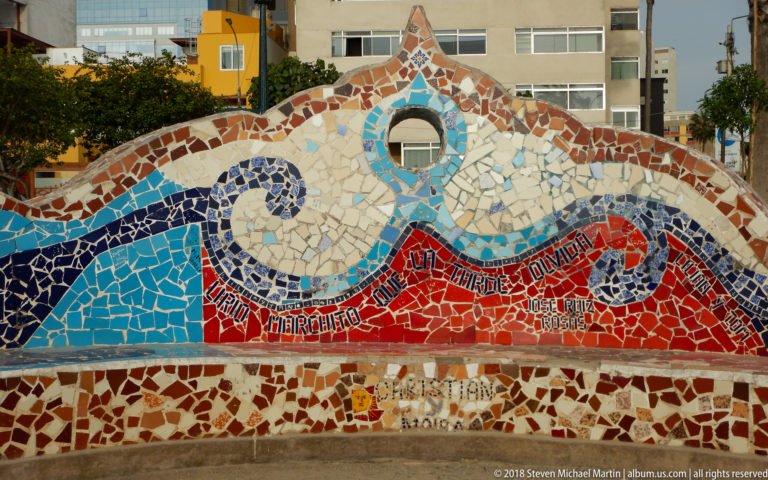 Peaked mosaic wall with blue-and-white wave spirals, red tile field, circular opening, and embedded names and inscriptions.
