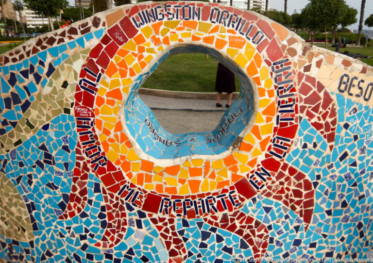 Circular opening in a colorful mosaic wall with Spanish words and names, framing lawn and a passerby beyond.