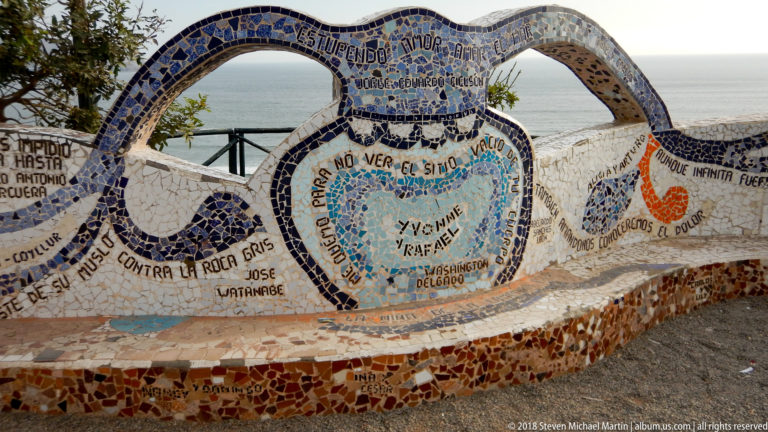 Blue-and-white mosaic bench with rounded openings, handwritten names and phrases, and the Pacific visible through the backrest.