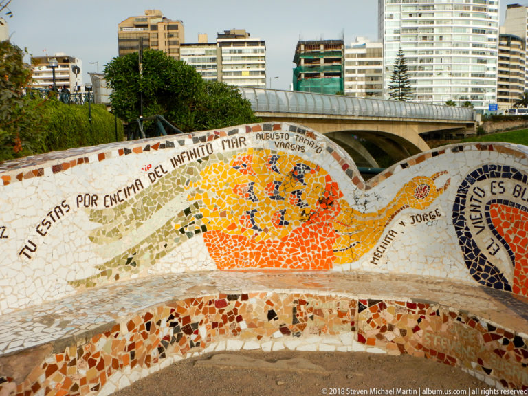 Curved mosaic wall with Spanish text and a large orange sun motif, with Miraflores apartment buildings behind it.
