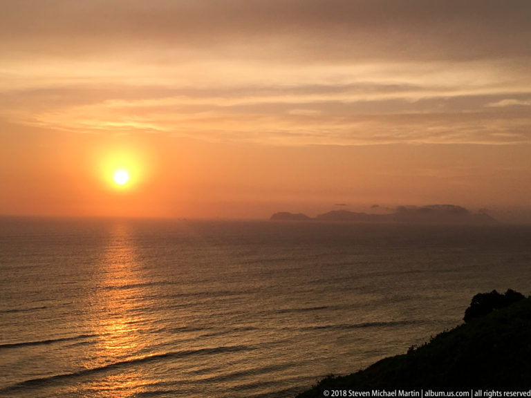 Bright orange sun over the Pacific with a long reflection, hazy low landforms on the horizon, and dark coastal vegetation.