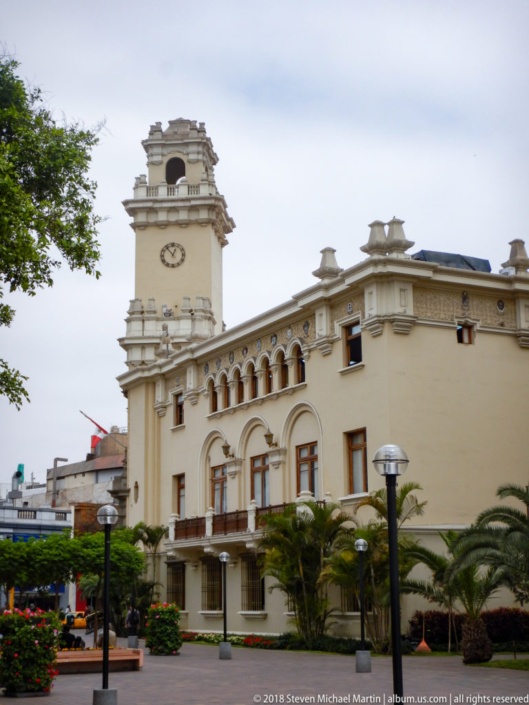 Pale municipal building with a tall central clock tower, arched windows, palms, and landscaped gardens in front.