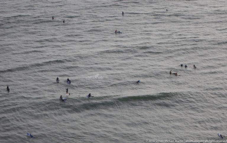 Numerous surfers on boards spread across gray Pacific swells below the Miraflores coast, with several small waves breaking.