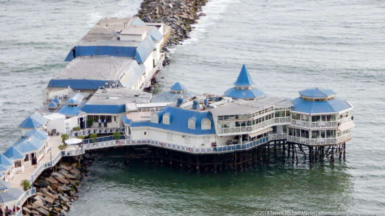 Close elevated view of La Rosa Náutica with blue roofs, glass-walled pavilions, pilings, and a narrow approach over the Pacific.