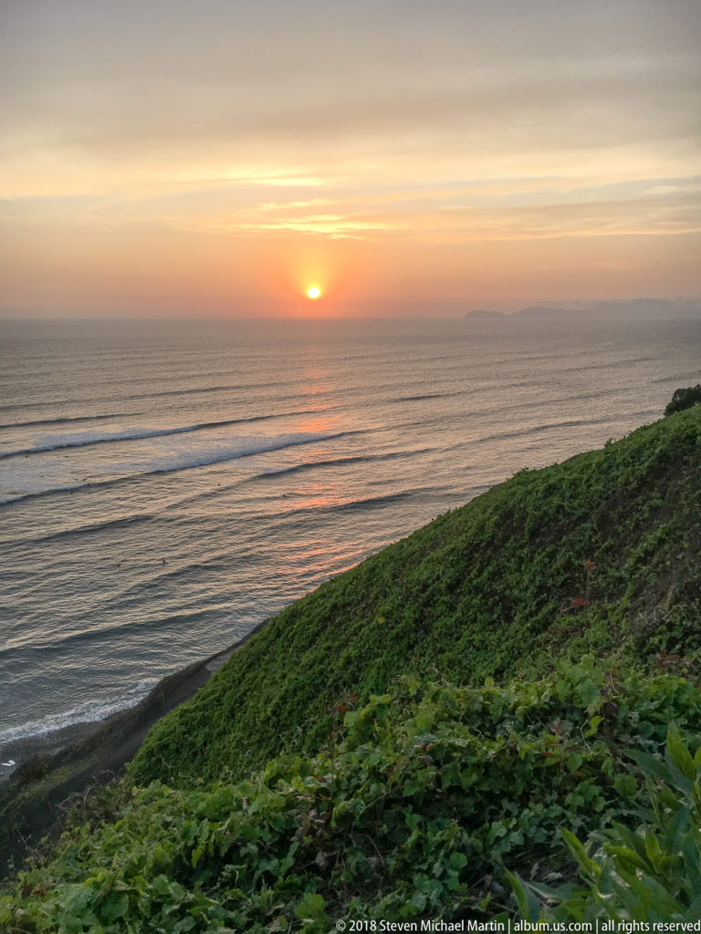 Orange sunset over the Pacific with a broad green Miraflores bluff descending across the foreground.