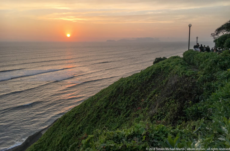 Sunset over the Pacific beside a steep green Miraflores cliff, with people gathered at a bluff-top lookout.