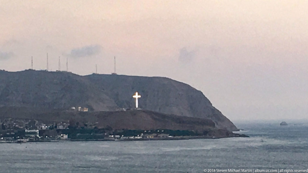 Distant Morro Solar headland across the bay, with a large white cross on the slope above shoreline buildings and the Pacific.