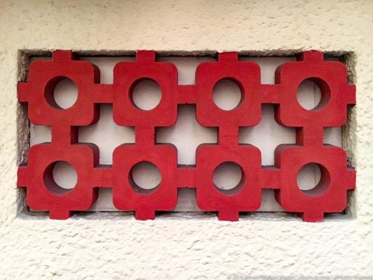 Red geometric concrete lattice with circular openings and cross-shaped gaps set into a textured white wall.