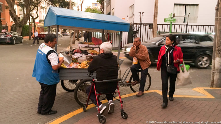 Street vendor at a blue-canopied cart serving pedestrians, including an older customer with a walker, on a Miraflores sidewalk.