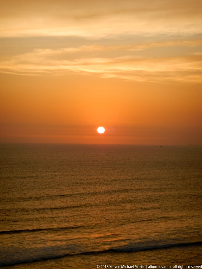 Low orange sun on the Pacific horizon beneath layered clouds, with dark parallel swells in the foreground.