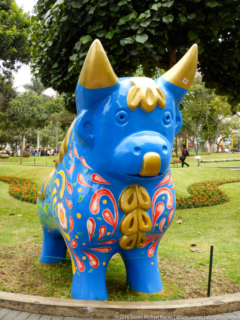 Large bright-blue bull sculpture with yellow horns and multicolored painted decoration standing on a park lawn.