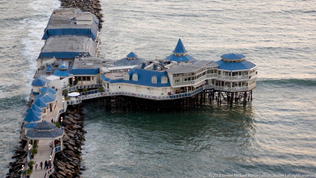 Elevated view of the blue-roofed La Rosa Náutica restaurant on pilings and a breakwater extending into the Pacific below Miraflores.