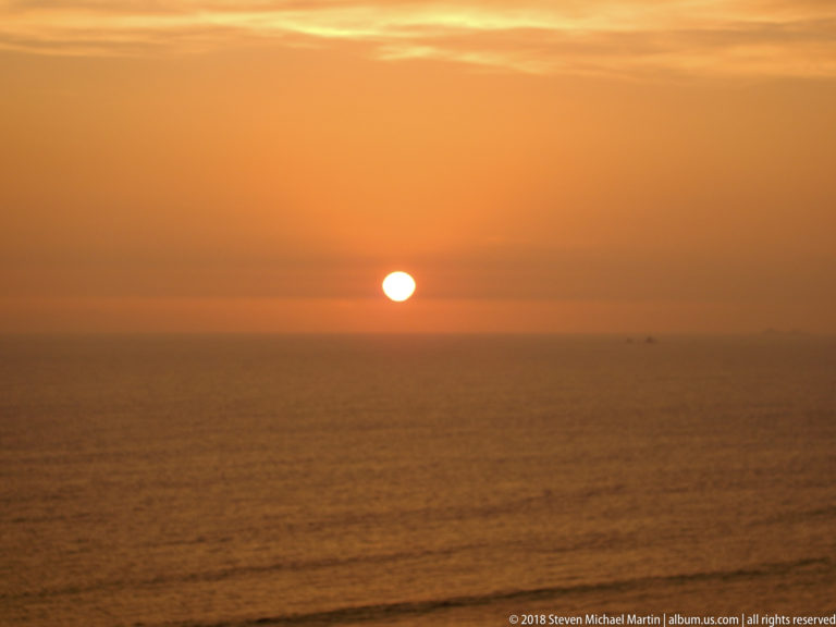 Orange sun just above the Pacific Ocean horizon, with hazy sky and calm dark water off Miraflores.