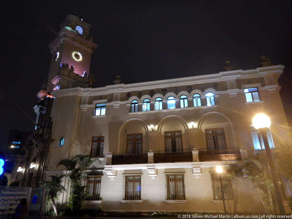 Illuminated Miraflores building with arched ground floor, balconies, upper windows, palms, and a tall corner clock tower at night.