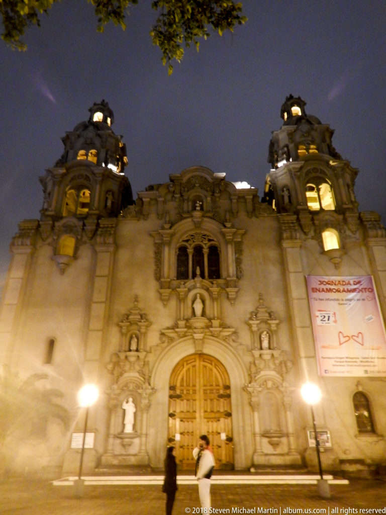 Upward night view of an ornate Miraflores façade framed by two illuminated domed towers and a dark blue sky.