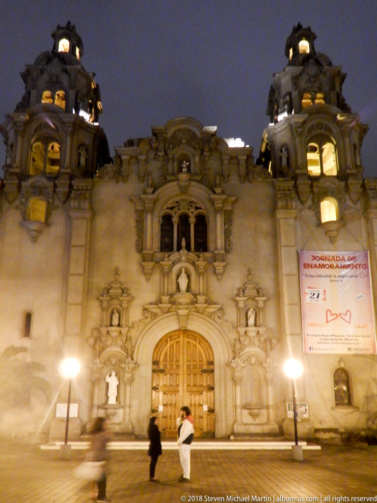 Frontal night view of an ornate Miraflores civic building with arched entrance, sculpted façade, twin domed towers, and two pedestrians.