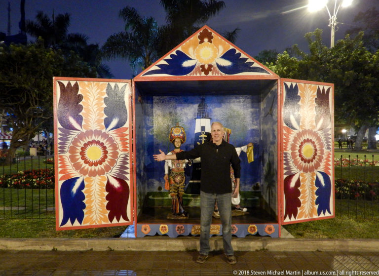 Man standing beside a colorful shrine-like park display with painted flowers, birds, and small figures in Miraflores.